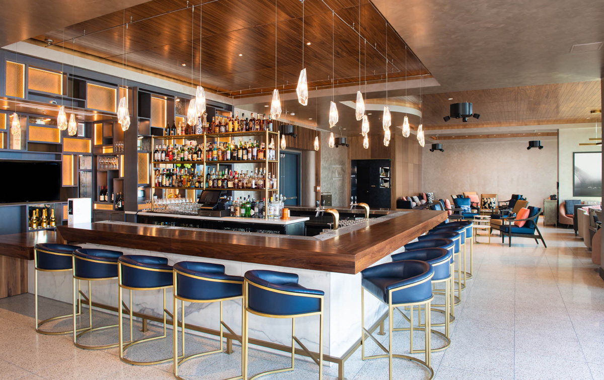 A modern bar with a long wooden counter, blue stools, and pendant lights.