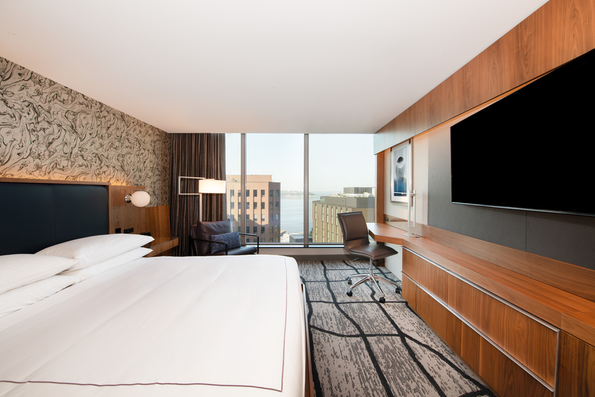 A hotel room with a bed, TV, desk, and window with a city view.